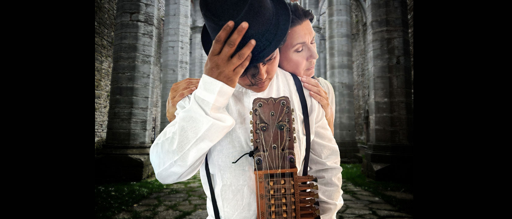 Två personer synns på bilden. Den främre håller i ett gammalt instrument och lutar sin svarta hatt över sitt ansikte. Den andra personen håller om den första personen bakifrån. De båda är klädda i vitt